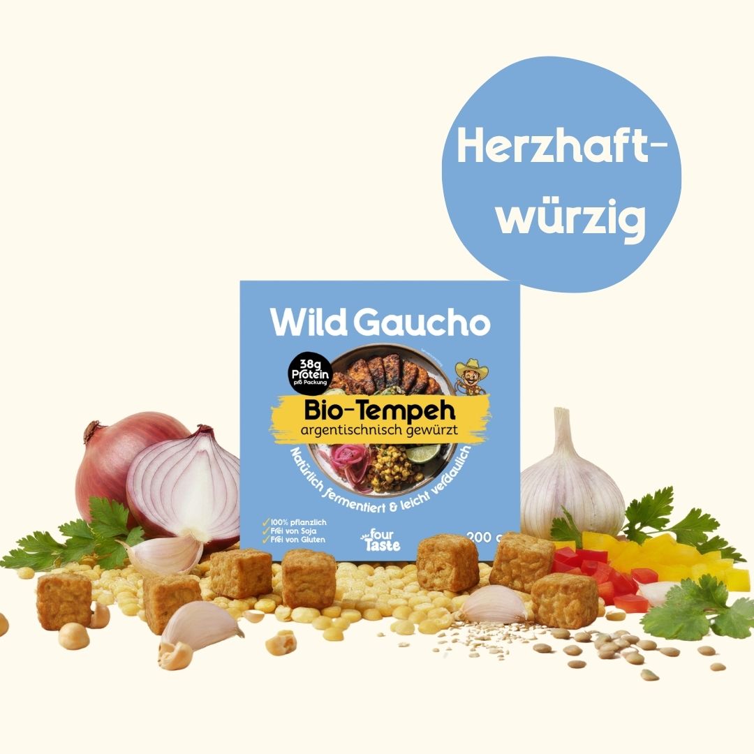 Eine Packung "Wild Gaucho" Bio-Tempeh in blauer Verpackung, angerichtet mit roten Zwiebeln und Knoblauch; ein Badge kennzeichnet den Geschmack als „herzhaft würzig“.