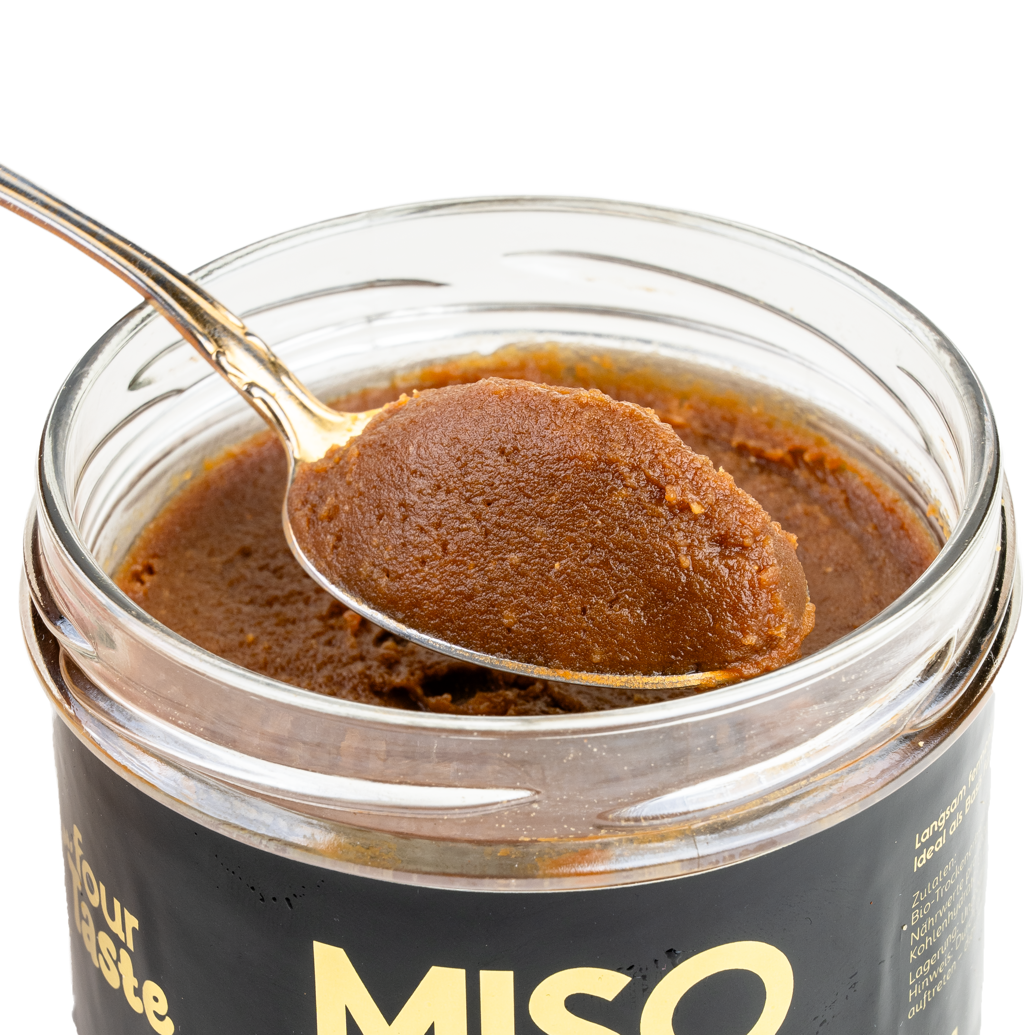 Ein goldener Löffel mit einer rotbraunen Miso Paste aus Erbsen die aus einem Glas mit schwarzem Etikett und goldener Schrift genommen wird von der Marke fourTaste